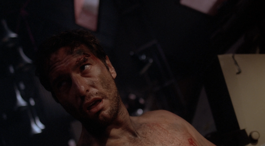 A shirtless, sweaty, visibly ill man leans back in a chair.
